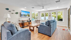 A modern living room with a wooden coffee table, two blue sofas, a TV mounted on the wall, and large windows offering a view of greenery outside. - Lazy Pelican 2 Bedroom Home by RedAwning (Holmes Beach)