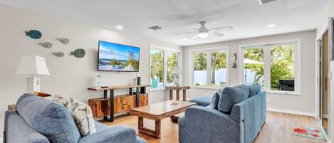 Ein modernes Wohnzimmer mit einem Holztisch, zwei blauen Sofas, einem an der Wand montierten Fernseher und großen Fenstern mit Blick ins Grüne.