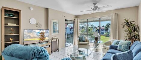 A spacious living room with a large TV, comfortable seating, a ceiling fan, and a view of the outdoors.