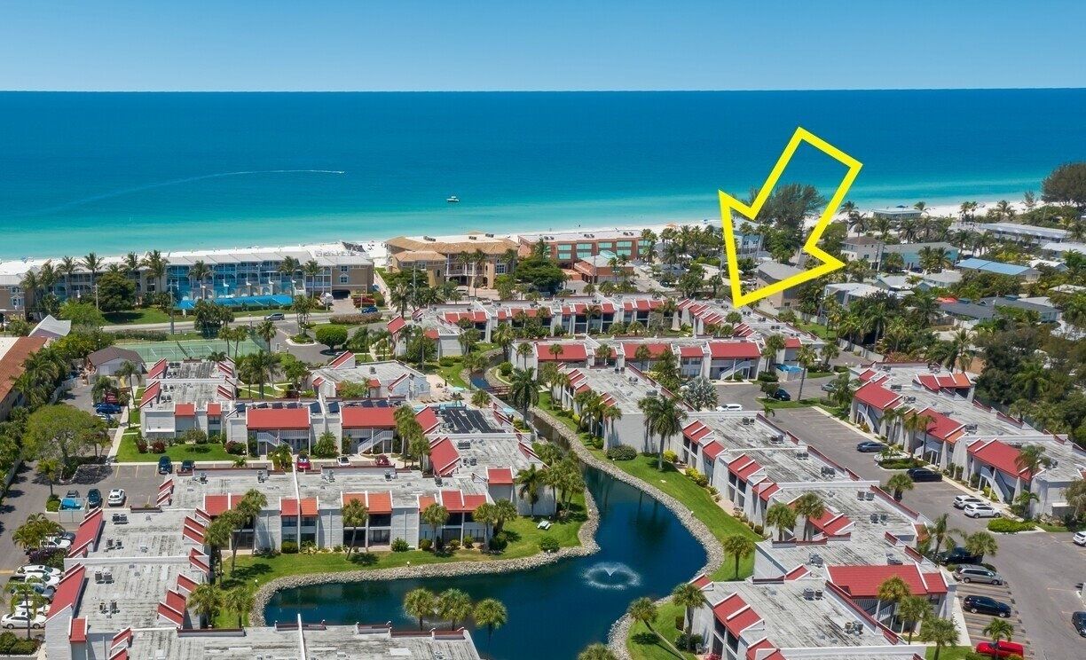 An aerial view of a resort with multiple buildings, a central water feature, and a beach in the background.