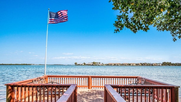 A wooden pier with a flagpole, leading to a body of water with a distant shoreline.