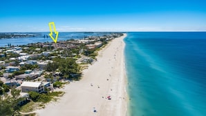 An aerial view of a coastal town with a long sandy beach, a pier extending into the water, and a mix of residential and commercial buildings along the shore.
