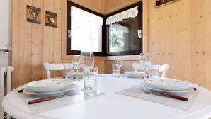 Mesa de comedor preparada para dos personas, con platos blancos, vasos transparentes y cubiertos. La habitación tiene paredes de madera y una ventana con cortinas blancas.