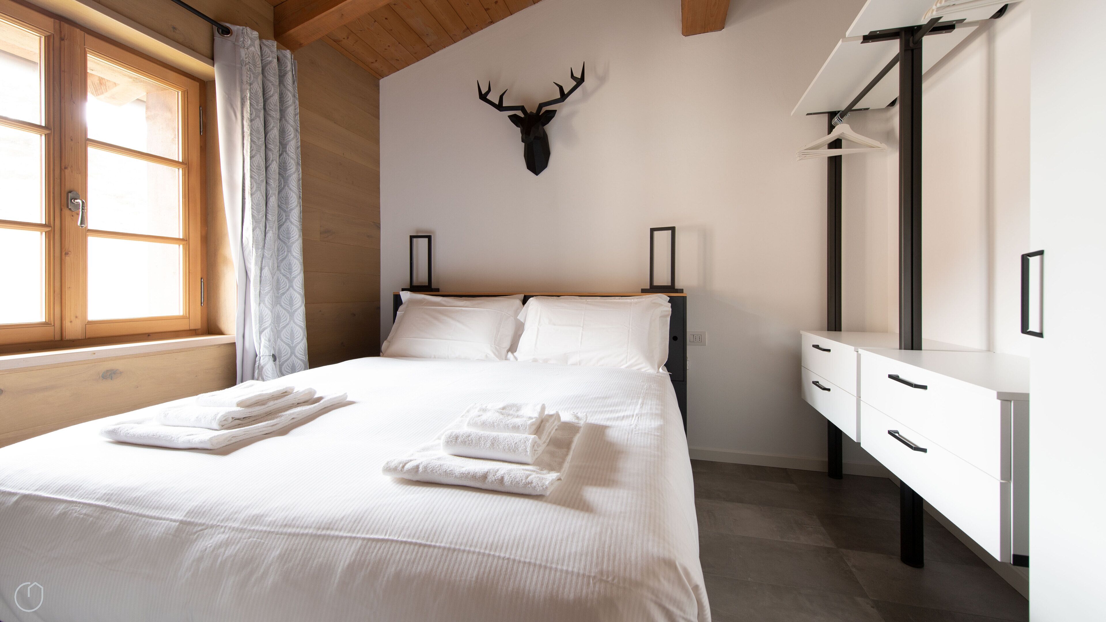 Une chambre d’hôtel avec un lit, du linge de lit blanc, une tête de cerf fixée au mur, une commode et une fenêtre avec des rideaux.