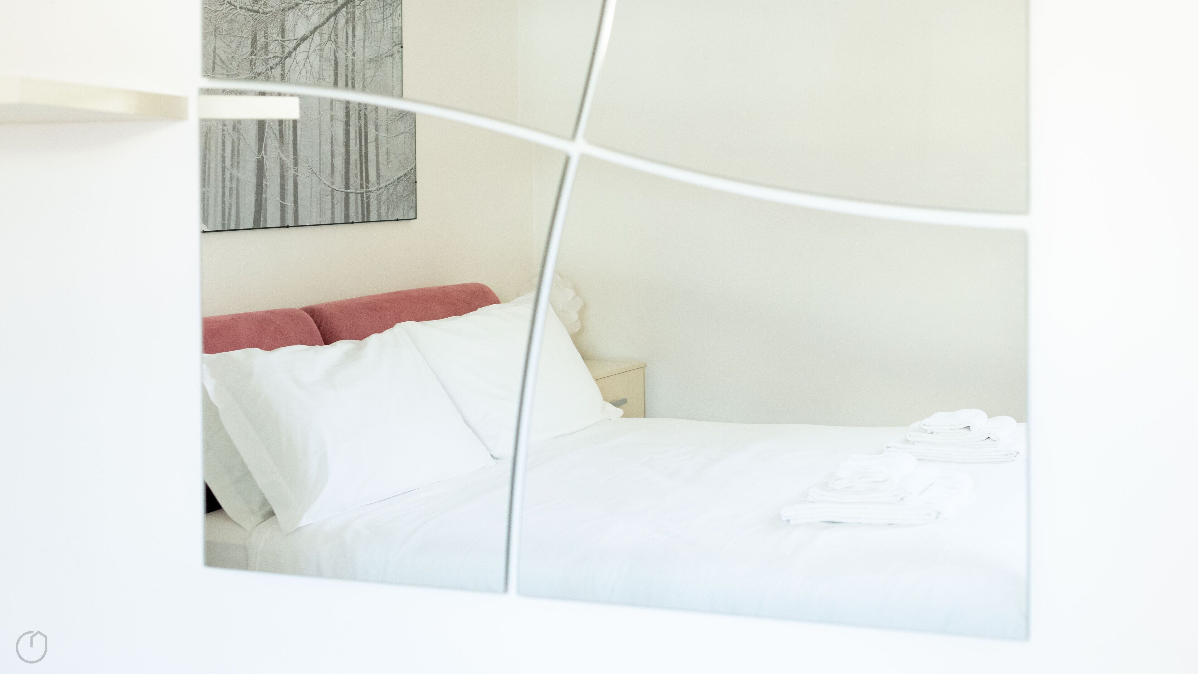 A neatly made bed with white linens and a red headboard, a small bedside table, and a wall-mounted artwork.