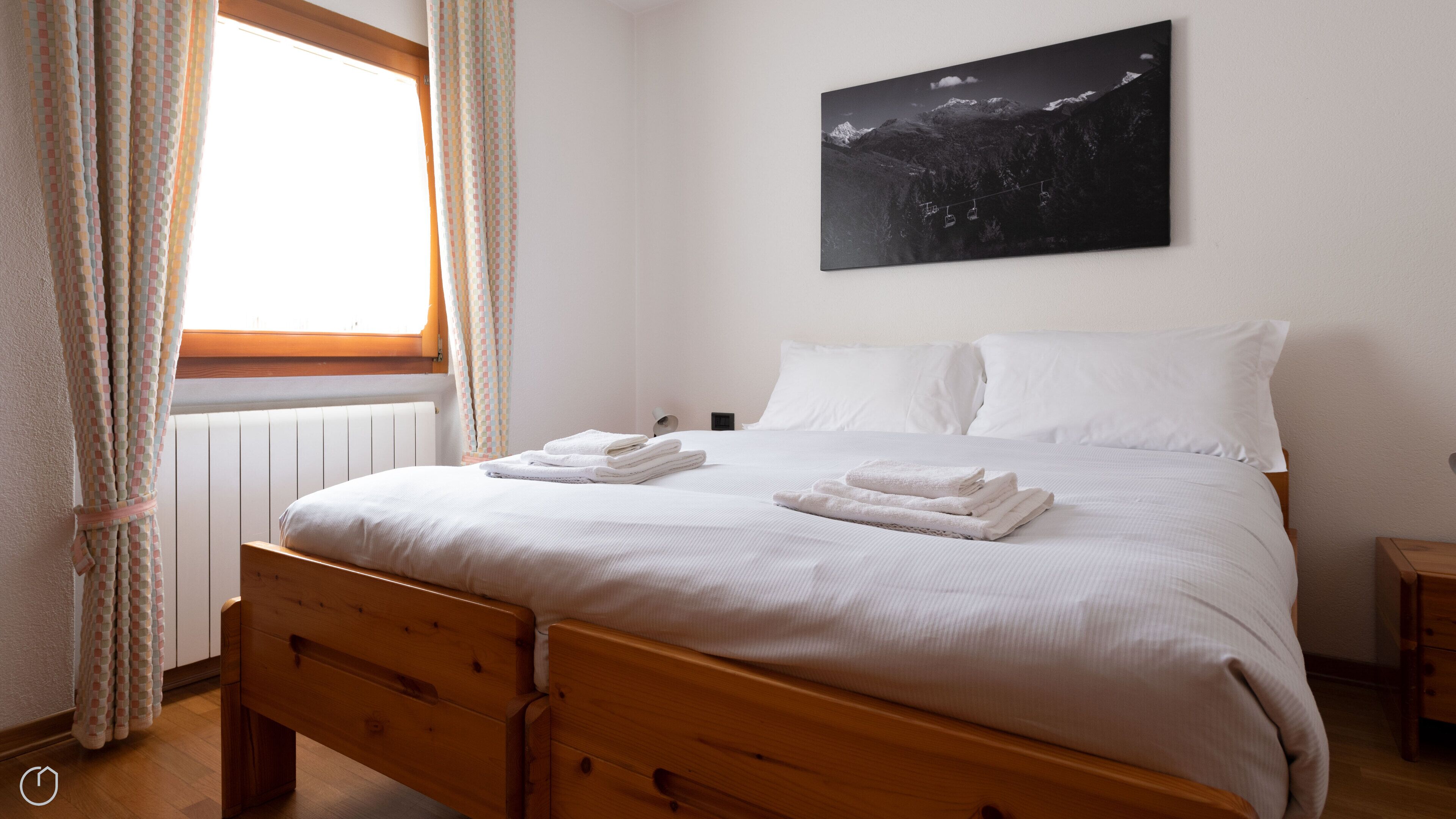 A neatly made bed with white linens and folded towels, a wooden headboard, a window with curtains, and a mountain landscape painting on the wall.