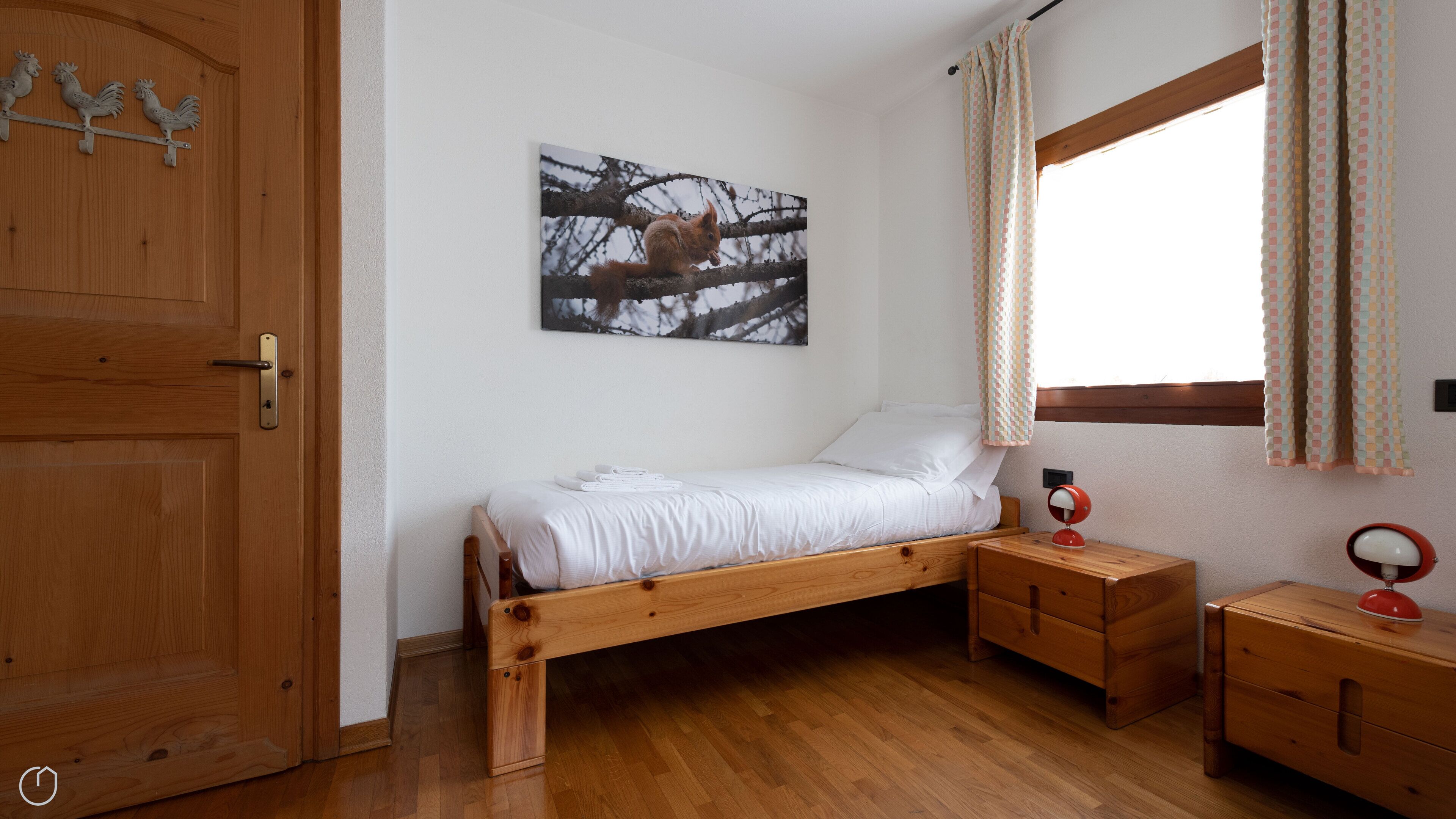 Ein Einzelzimmer mit einem hölzernen Bett, einem Nachttisch, einem kleinen Tisch mit Lampe, einem Fenster mit Vorhängen und einem Wandbild.