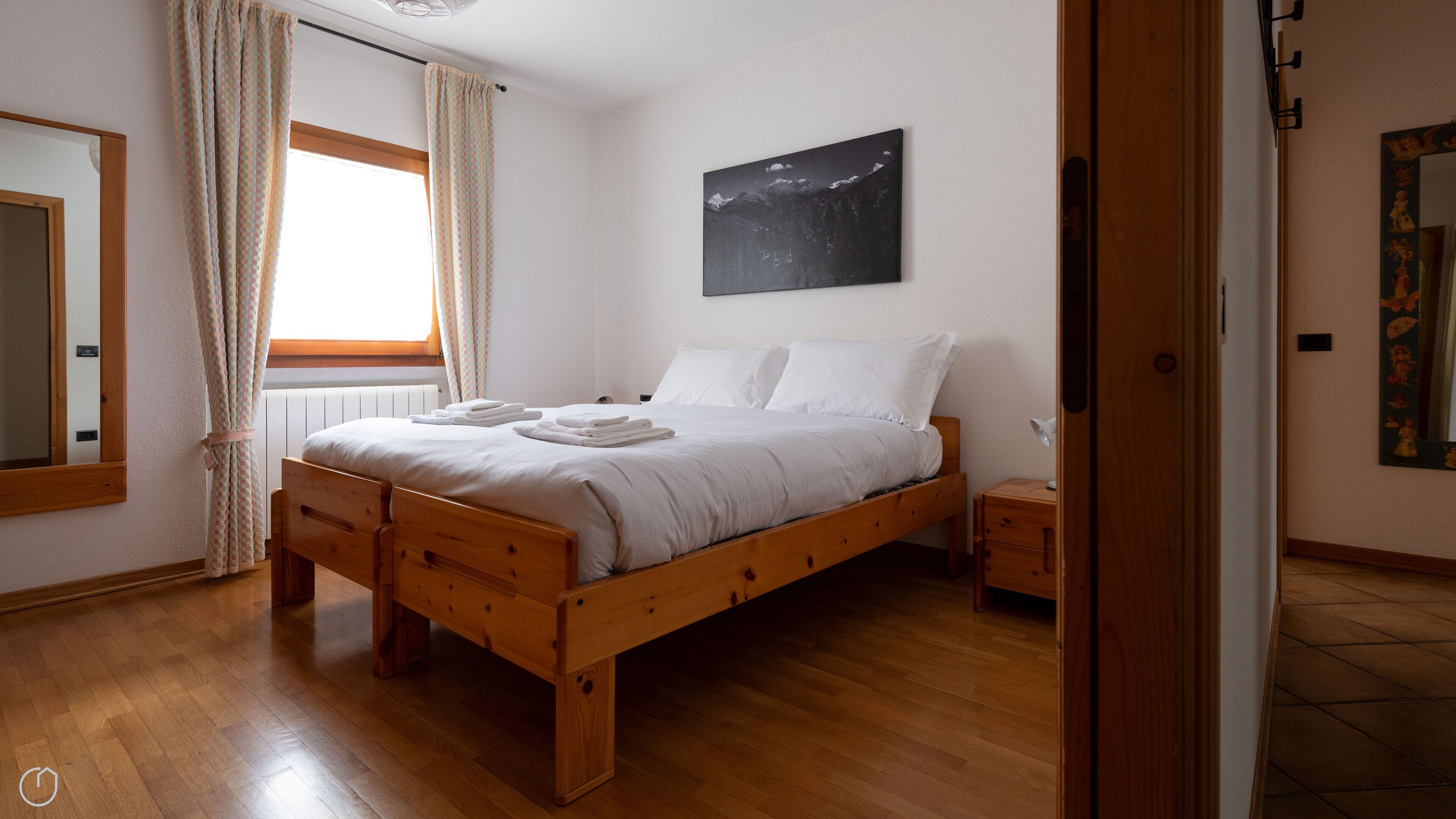 Un dormitorio con una cama de madera, una ventana con cortinas, una mesita de noche con lámpara y un cuadro colgado en la pared.