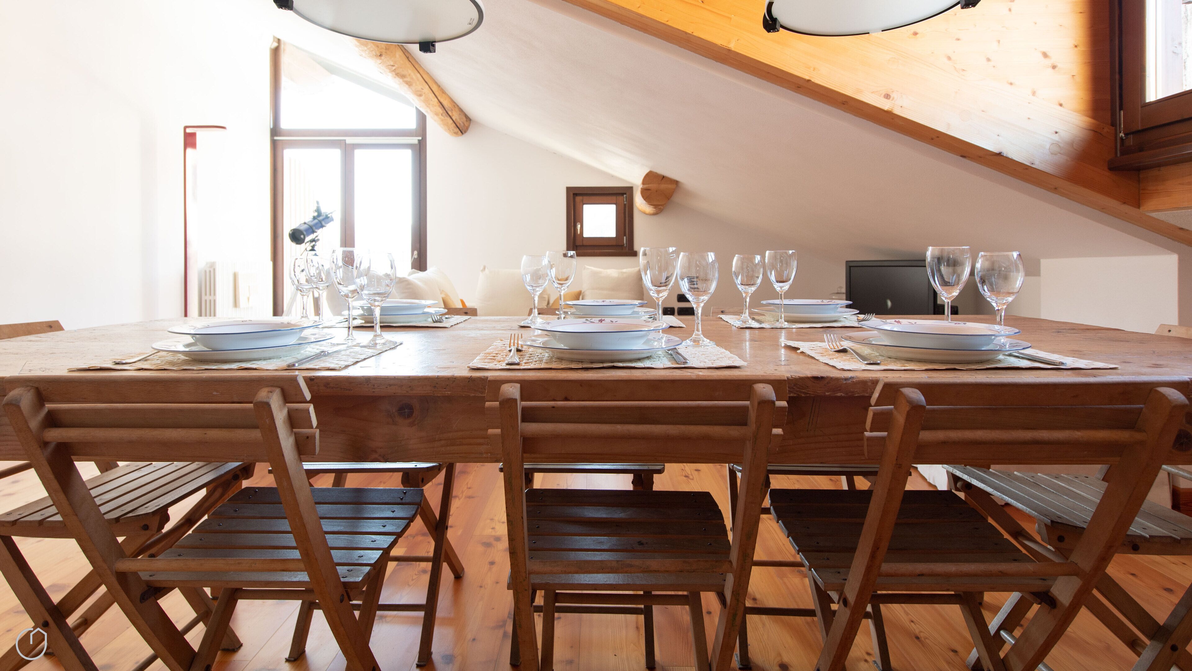 Une table à manger dressée avec des assiettes, des verres et des couverts, entourée de chaises en bois dans une pièce aux poutres apparentes et dotée d’une fenêtre.