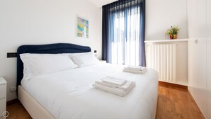 A neatly made bed with white linens and folded towels, a dark blue headboard, a window with blue curtains, a small potted plant on a white shelf, and a radiator on the wall. - Sertorelli 2 (Bormio)