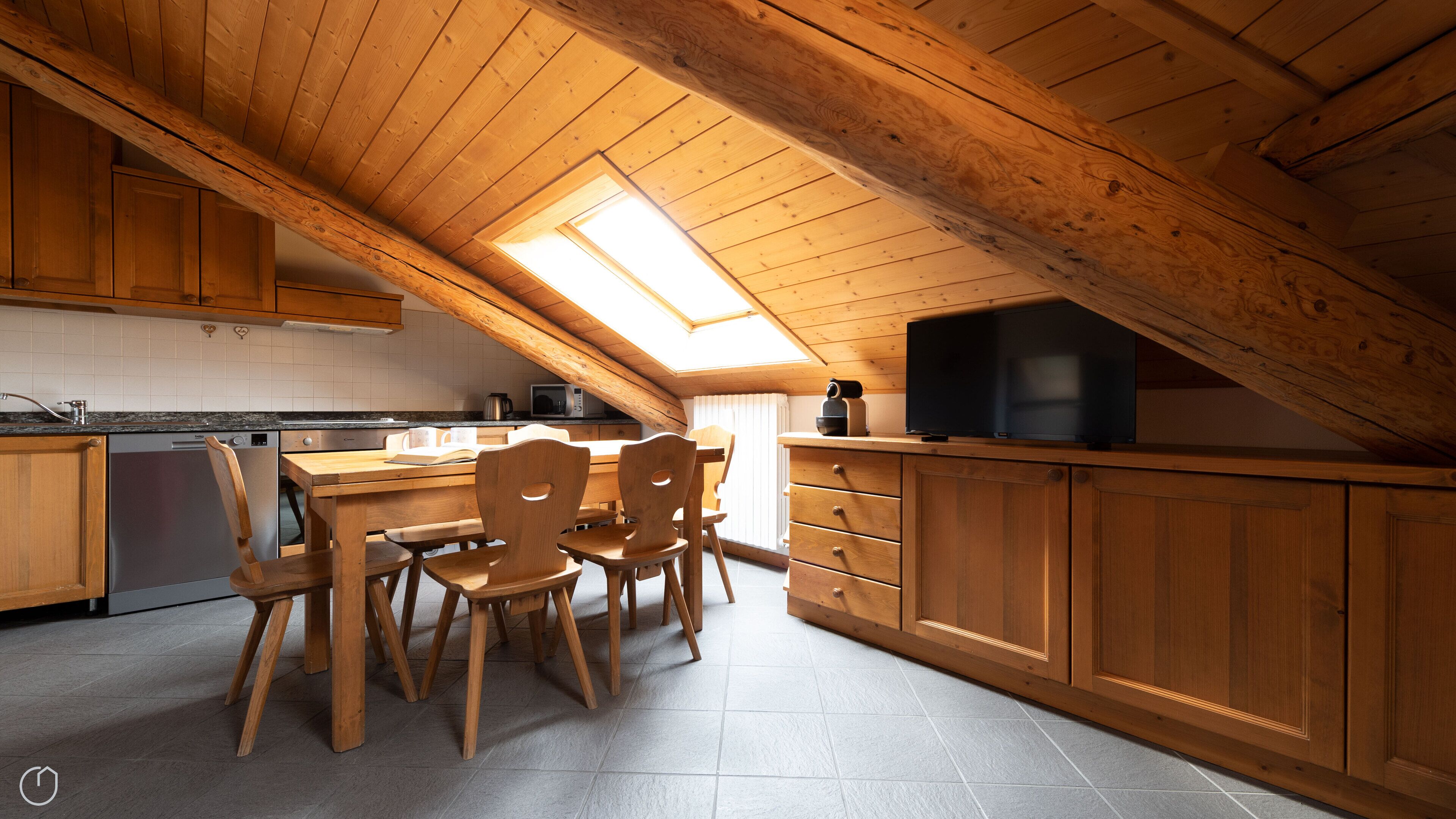 A kitchen with wooden cabinets, a dining table with chairs, a built-in oven, and a television on a wooden shelf.