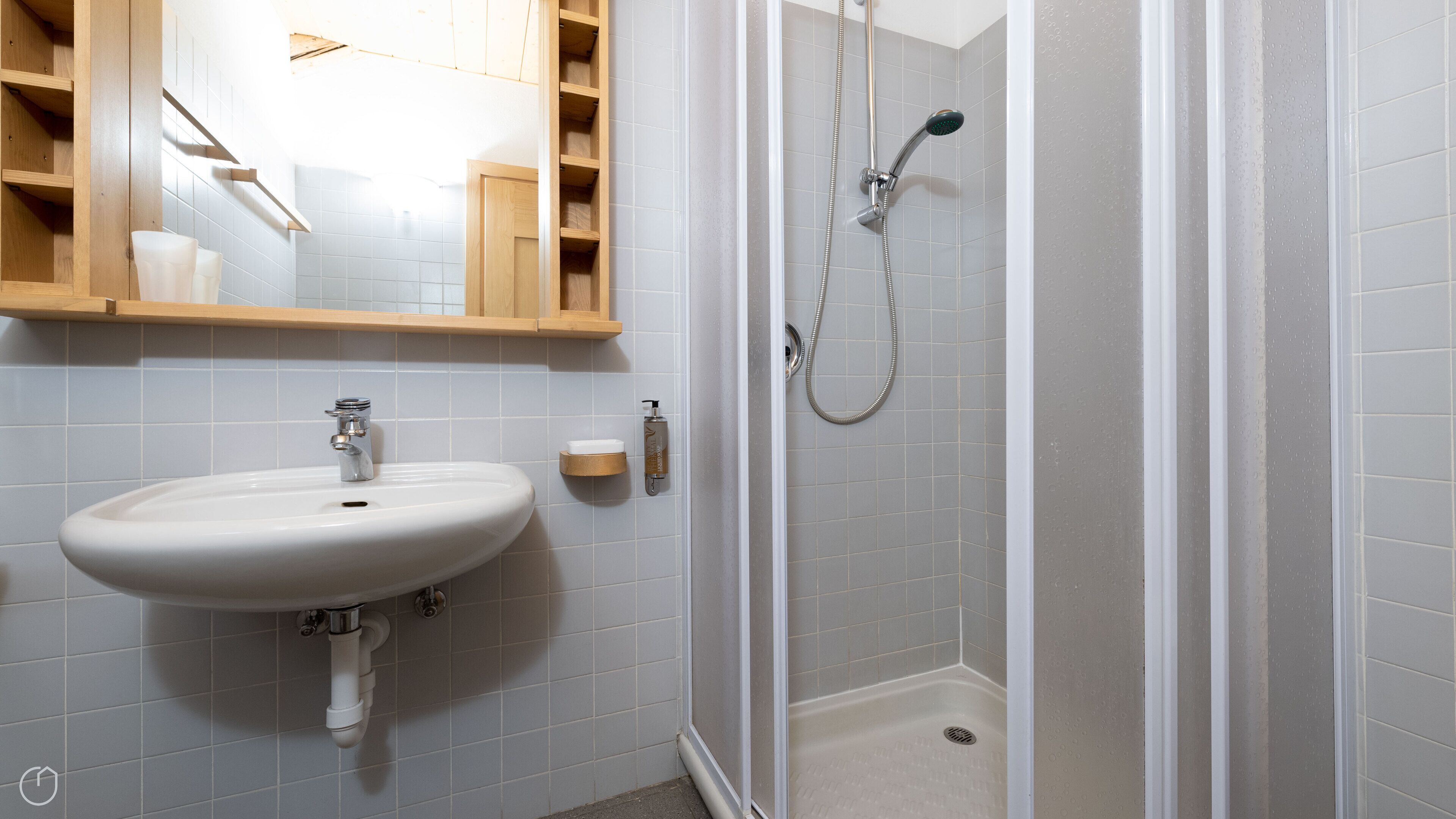 A bathroom with a white sink, a shower, and a wooden shelf above the sink.
