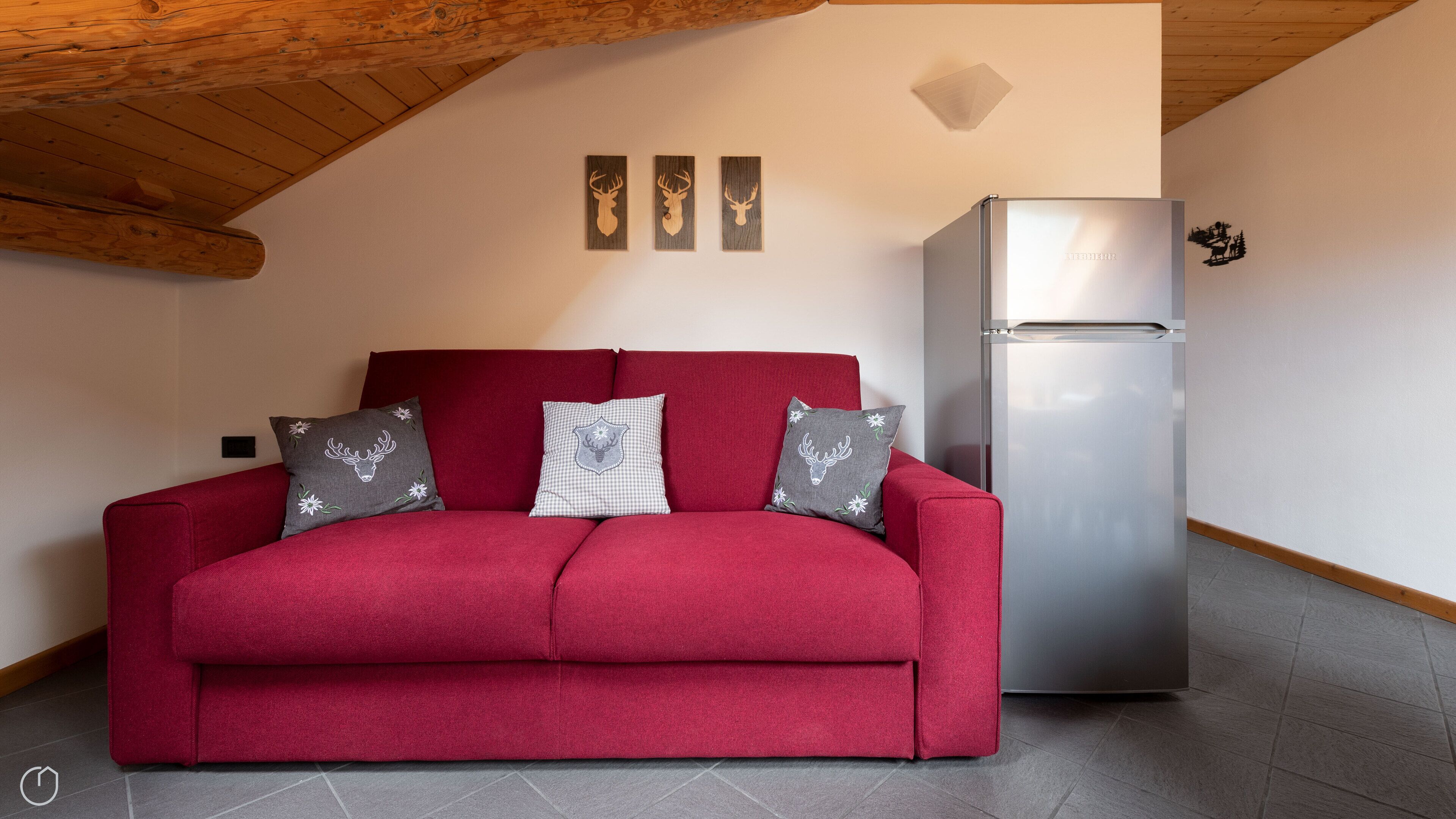 Un sofá rojo con tres cojines decorativos, un refrigerador de acero inoxidable y tres elementos decorativos de madera en la pared.
