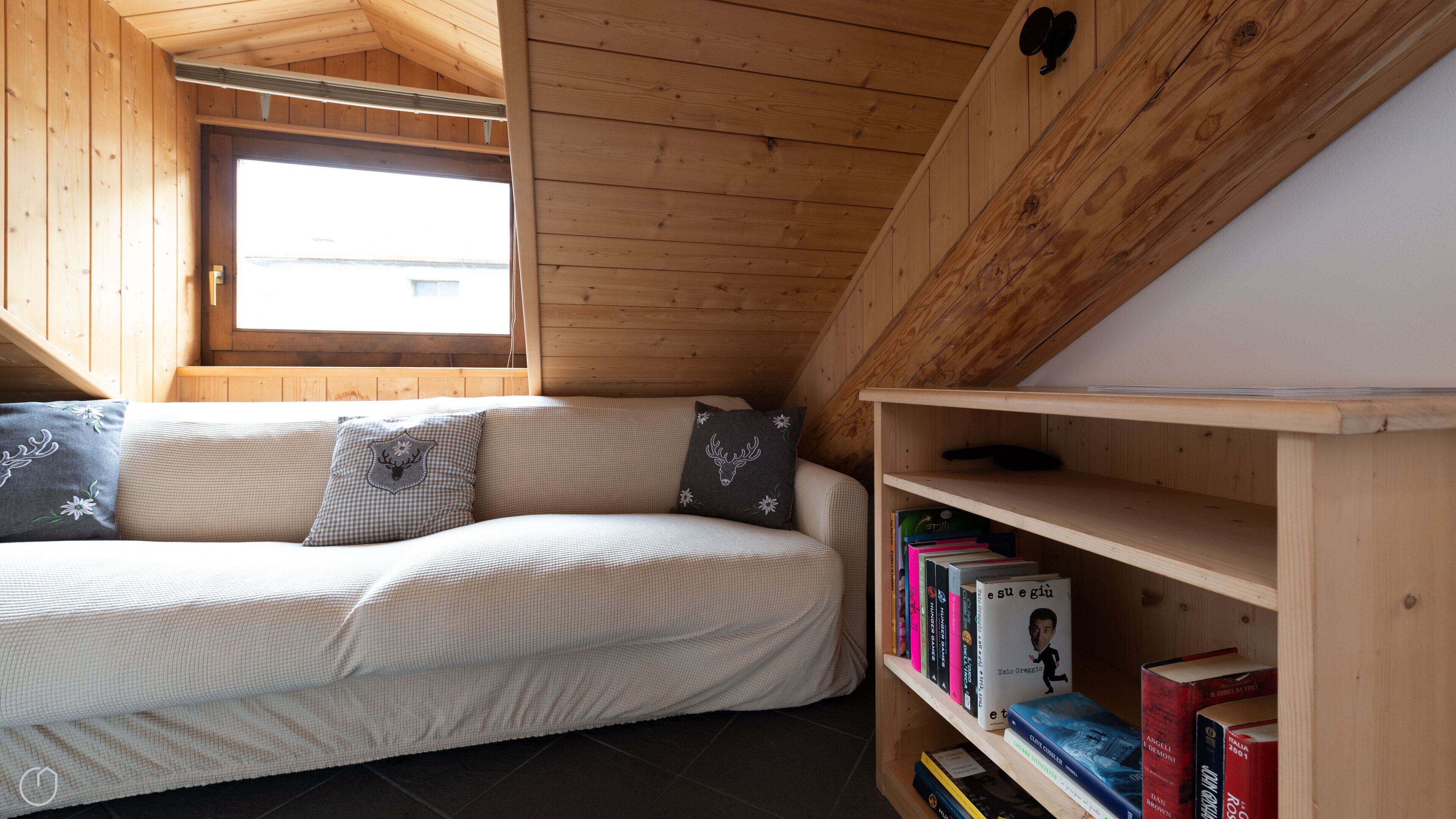 A cozy room with a wooden ceiling, a window, a sofa with patterned cushions, and a bookshelf with books.