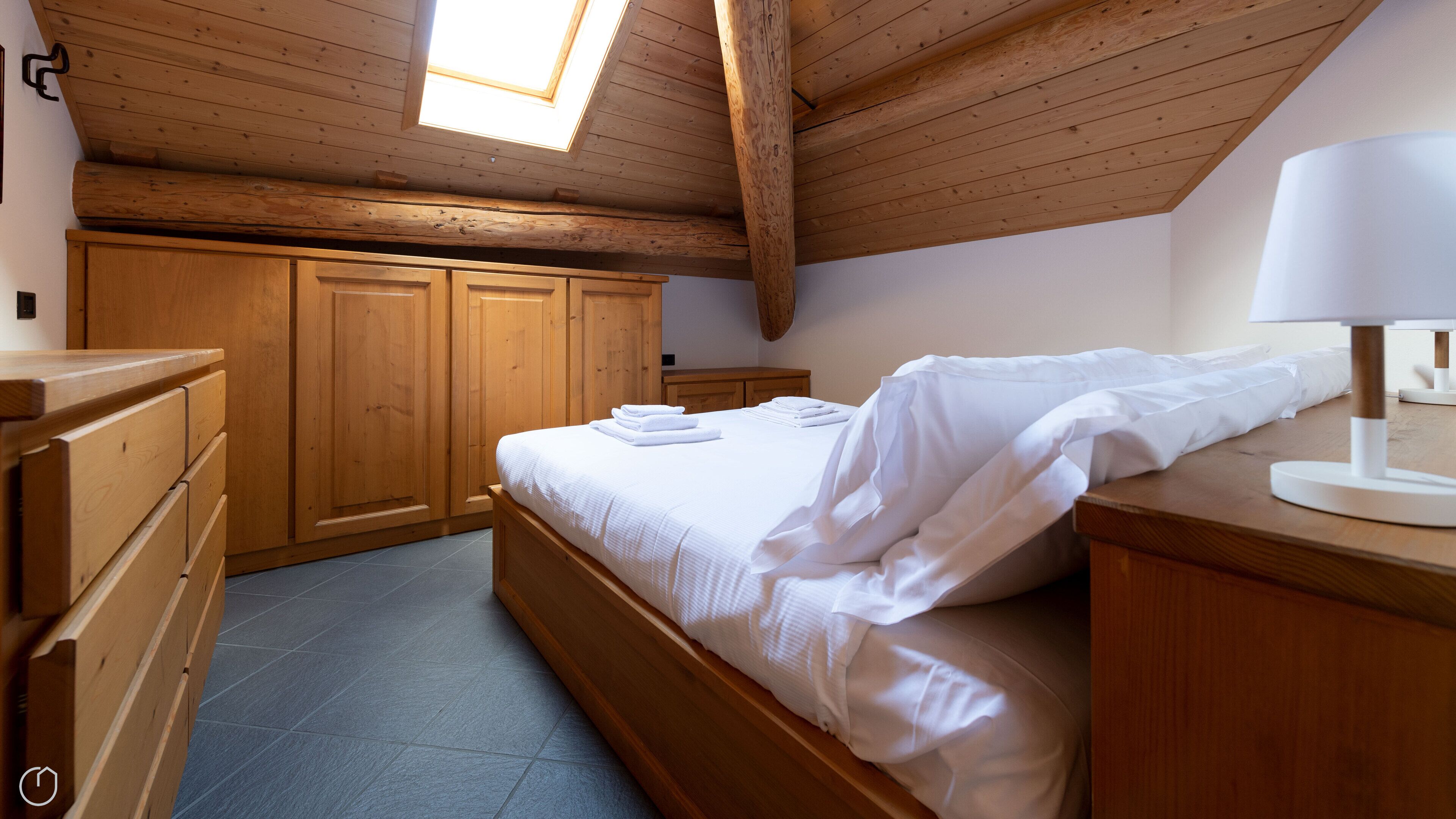 Ein Schlafzimmer mit Holzbalkendecke, Dachfenster, einem Bett mit weißen Leinen, einer Holzkommode und einem Nachttisch mit Lampe.