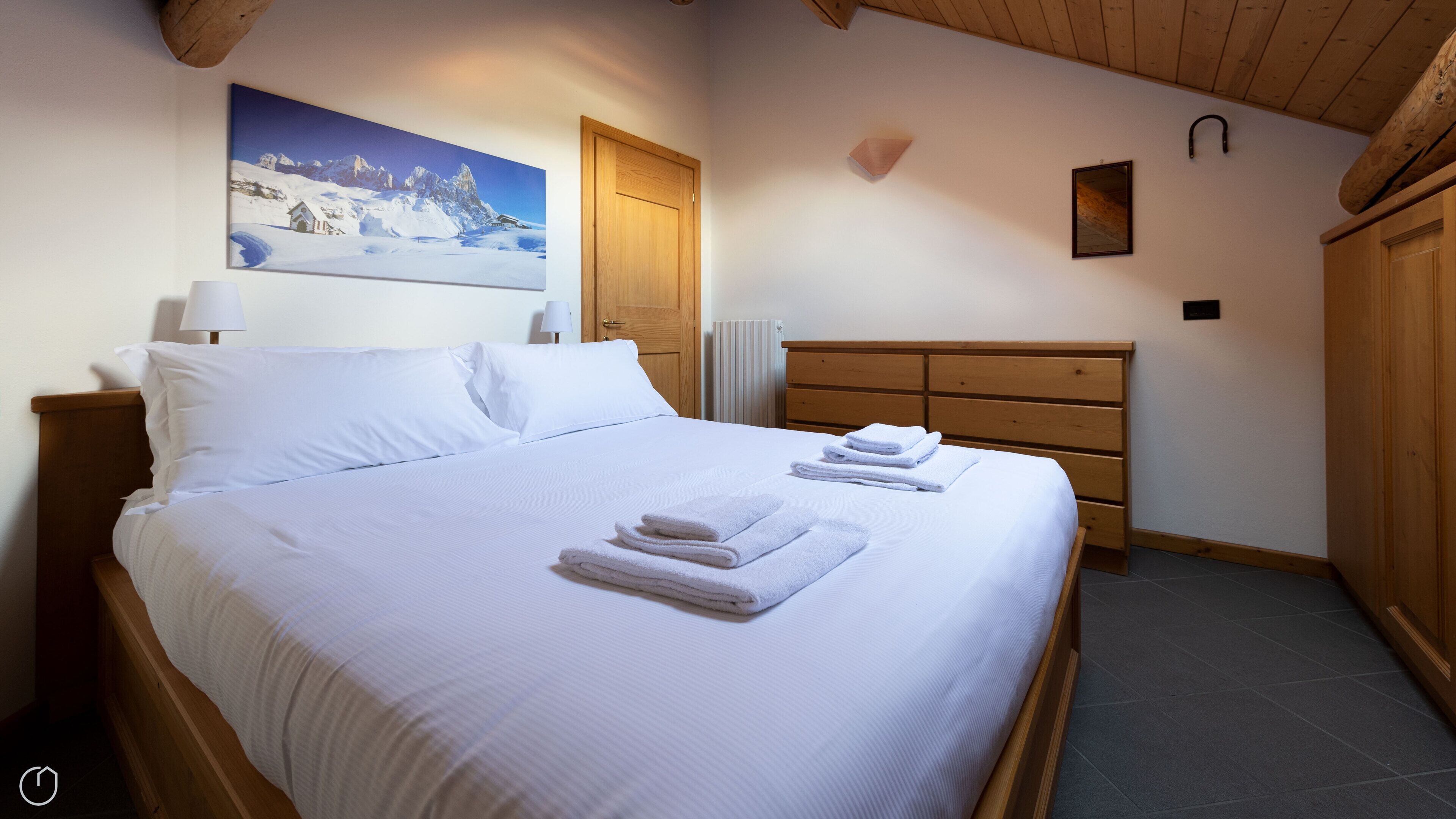 A neatly made bed with white linens and towels, a wooden headboard, a wooden dresser, and a painting of a snowy mountain landscape on the wall.