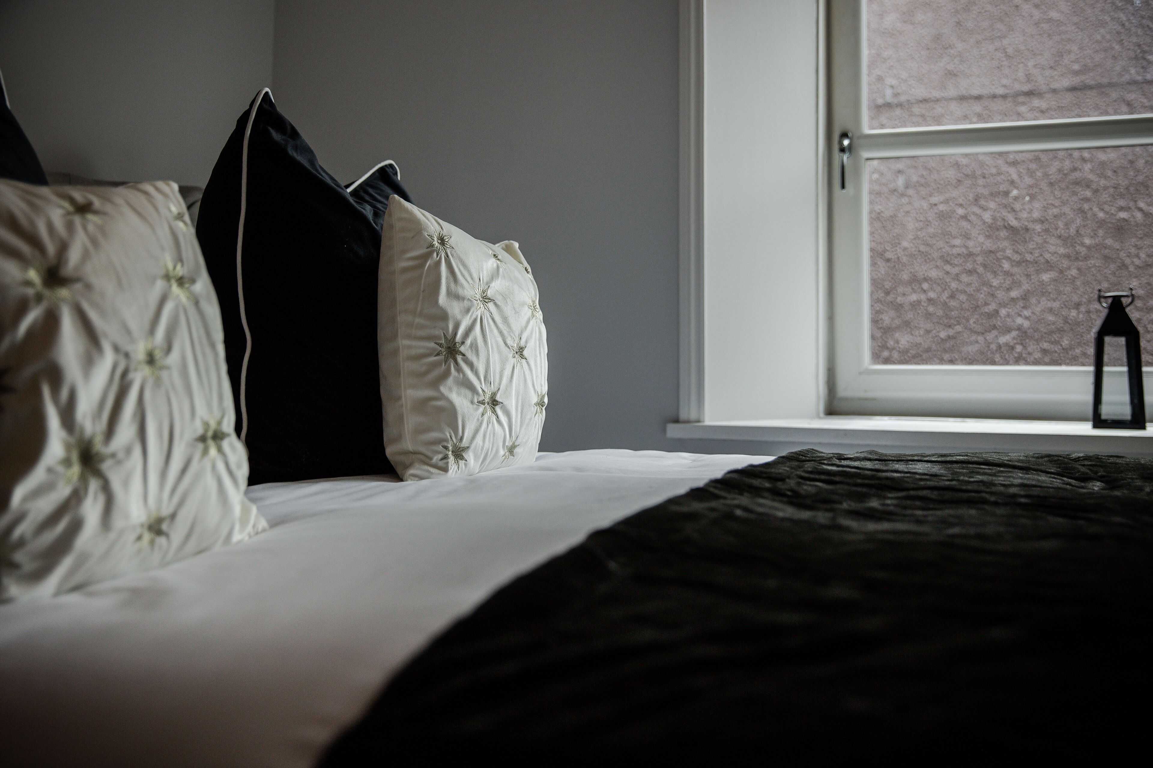 Cama bem arrumada com roupa de cama branca e preta, janela com vista para um edifício e porta à direita.