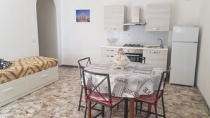 A compact living space with a kitchenette, a dining area with a table and chairs, and a bed with patterned bedding. - Apartment among ancient olive trees a short distance from the excavations of Velia. Sea view (Ascea)