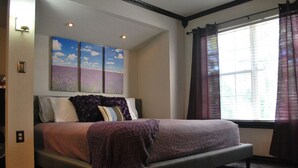 A bedroom with a bed, a window with curtains, and a painting above the bed.