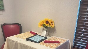 A dining table with a white tablecloth, red chairs, a tablet, a vase with sunflowers, and a small sign. - English Room at Whispering Pines Inn (Norman)