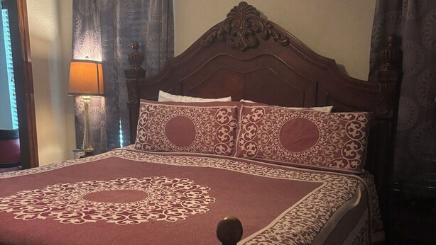 A neatly made bed with a patterned bedspread and pillows, a wooden headboard, and a bedside lamp. - English Room at Whispering Pines Inn (Norman)