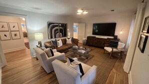 A living room with a brown leather sofa, white armchairs, a glass coffee table, and a large flat-screen TV. - Luxury Home - Walk to Grove and Square - Outdoor Patio Seating (Oxford)