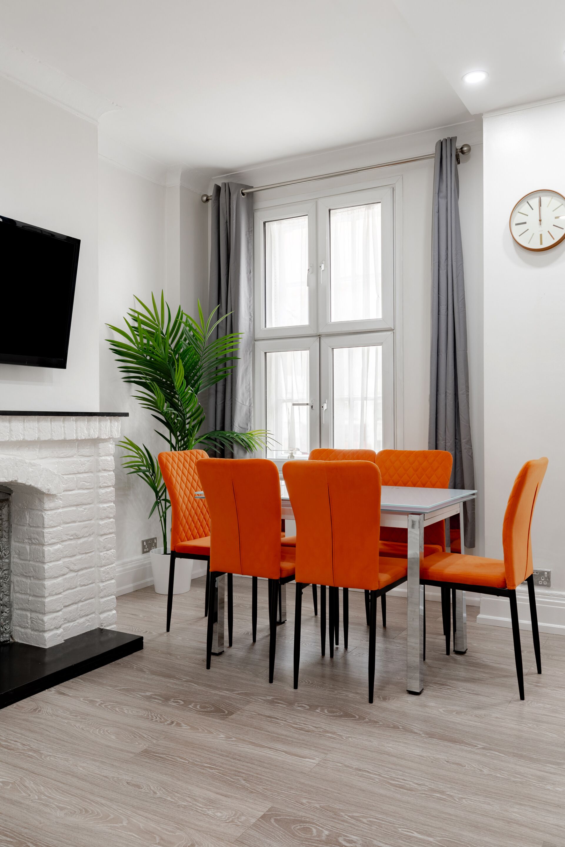A dining area with a white table and orange chairs, a white brick fireplace, and a large window with grey curtains.
