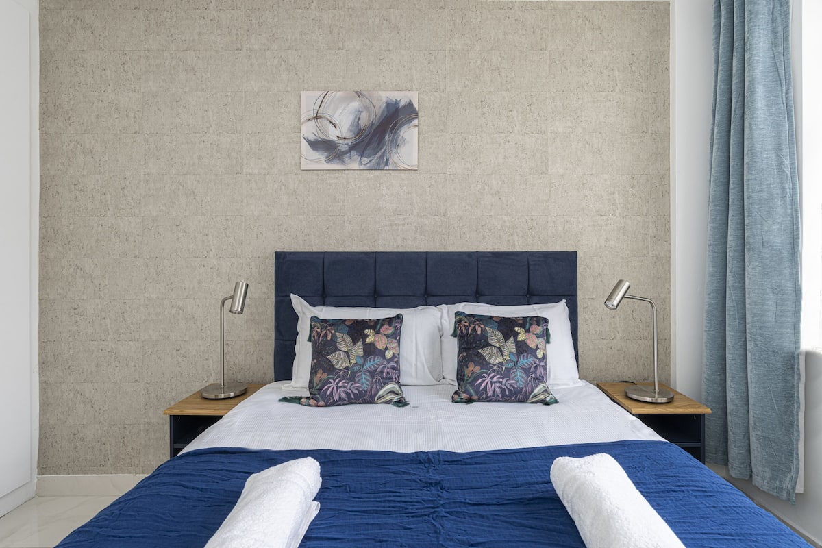 Una cama bien tendida con ropa de cama azul y blanca, dos almohadas decorativas, un cabecero y una obra de arte colgada en la pared.