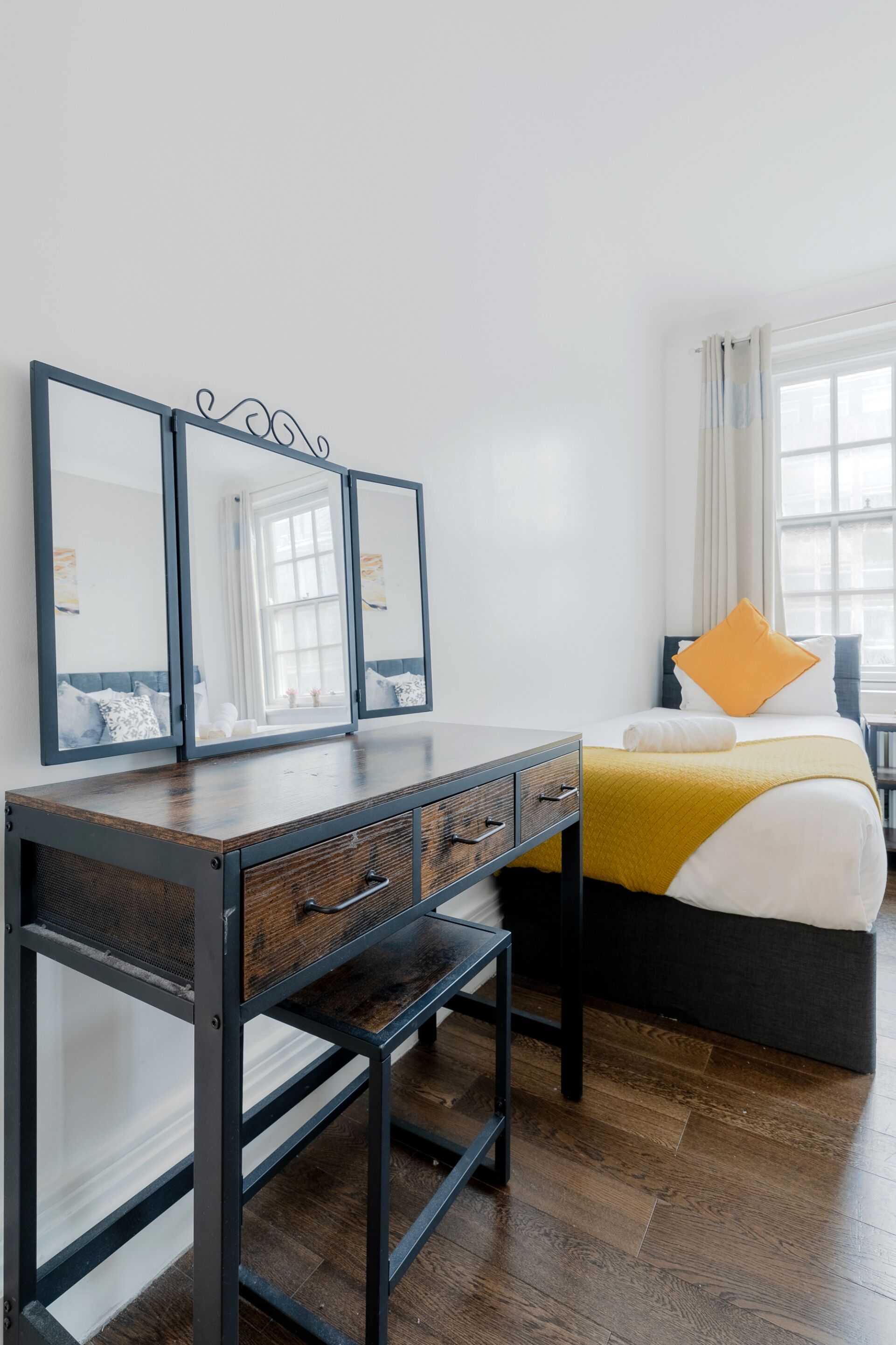 A hotel room with a wooden desk, a bed with a yellow blanket, and a large window.