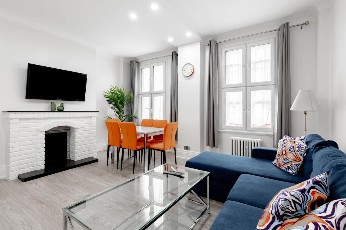 Una sala moderna con chimenea, televisor de pantalla plana, comedor con sillas naranjas y sofá azul con almohadones estampados.