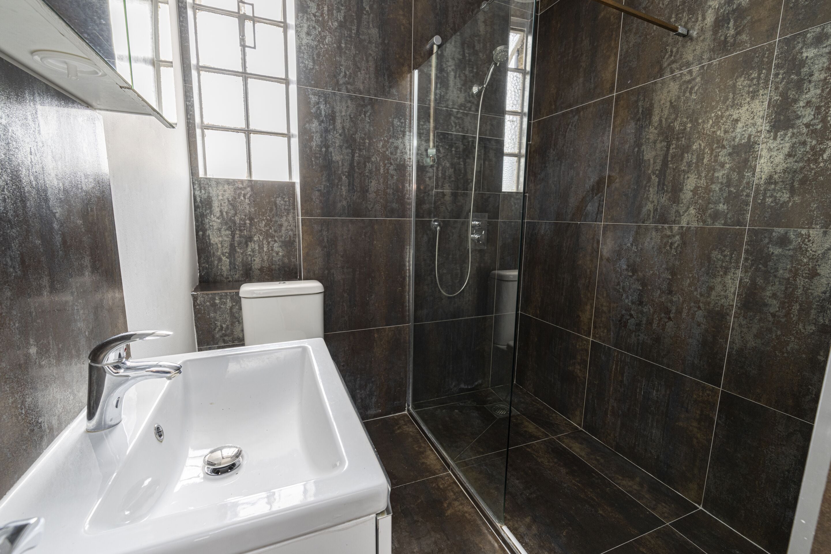 A bathroom with a white sink, a shower, and dark tiled walls.