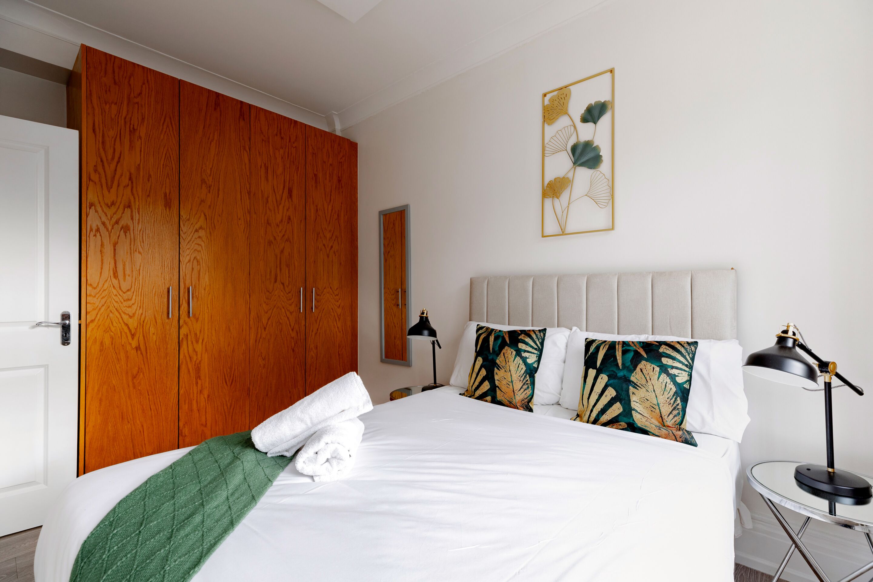 Una cama bien hecha con sábanas blancas y un plaid verde, un armario de madera y una obra de arte colgada en la pared.