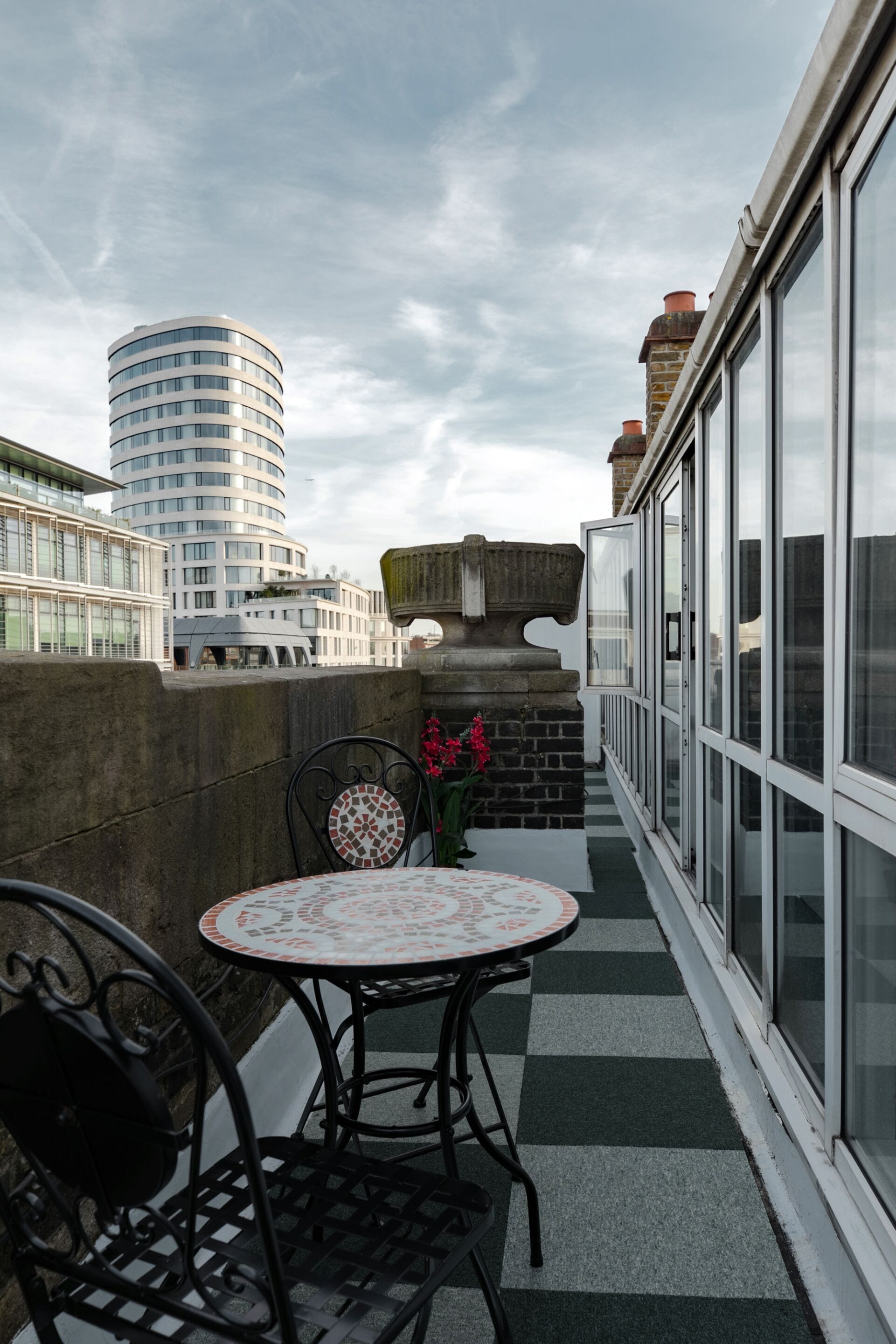 A balcony with a checkered floor, a round table with chairs, and a potted plant with flowers.