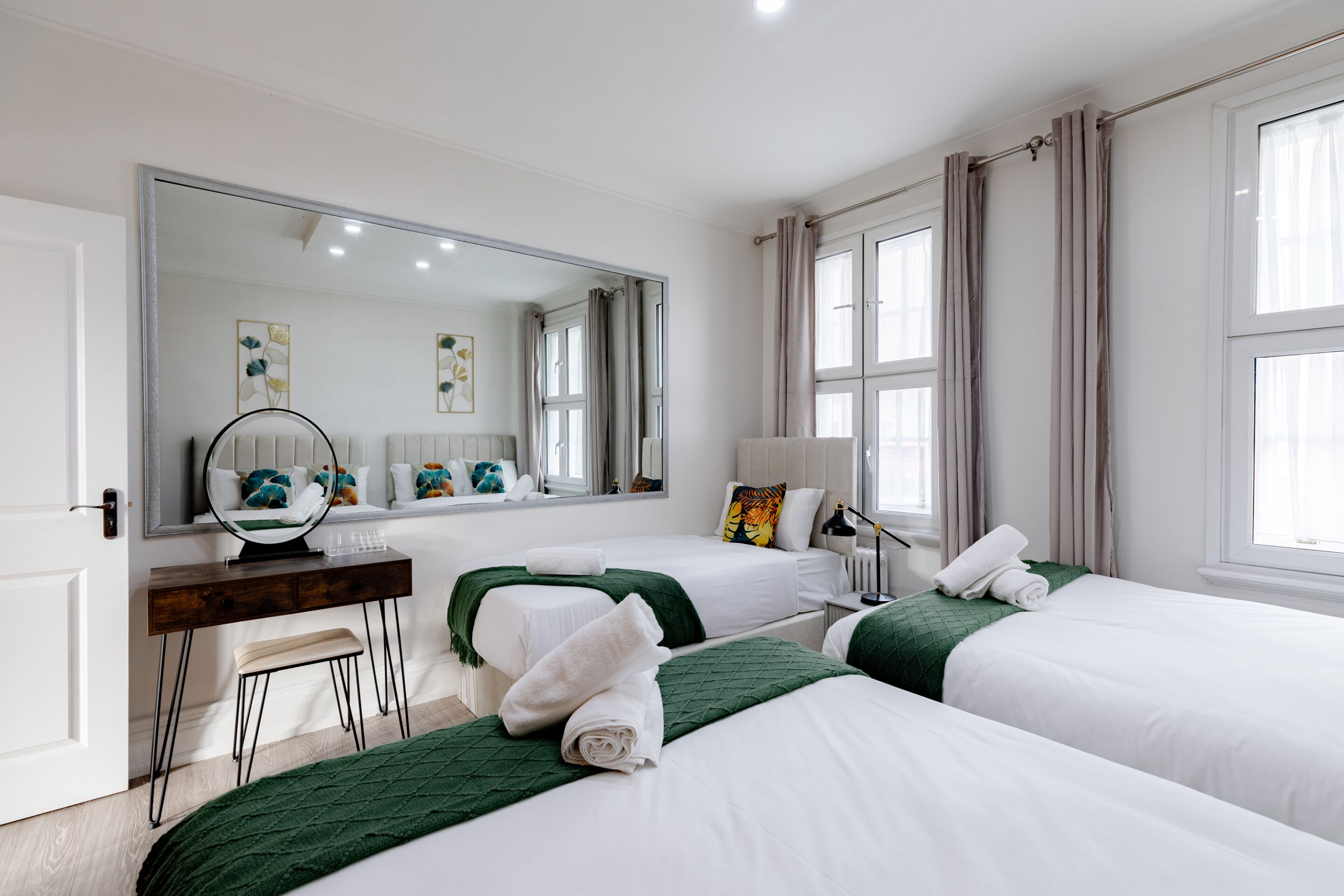 Ein Hotelzimmer mit zwei Betten, einem Schreibtisch, einem Stuhl, einem Spiegel und einem Fenster mit Vorhängen.