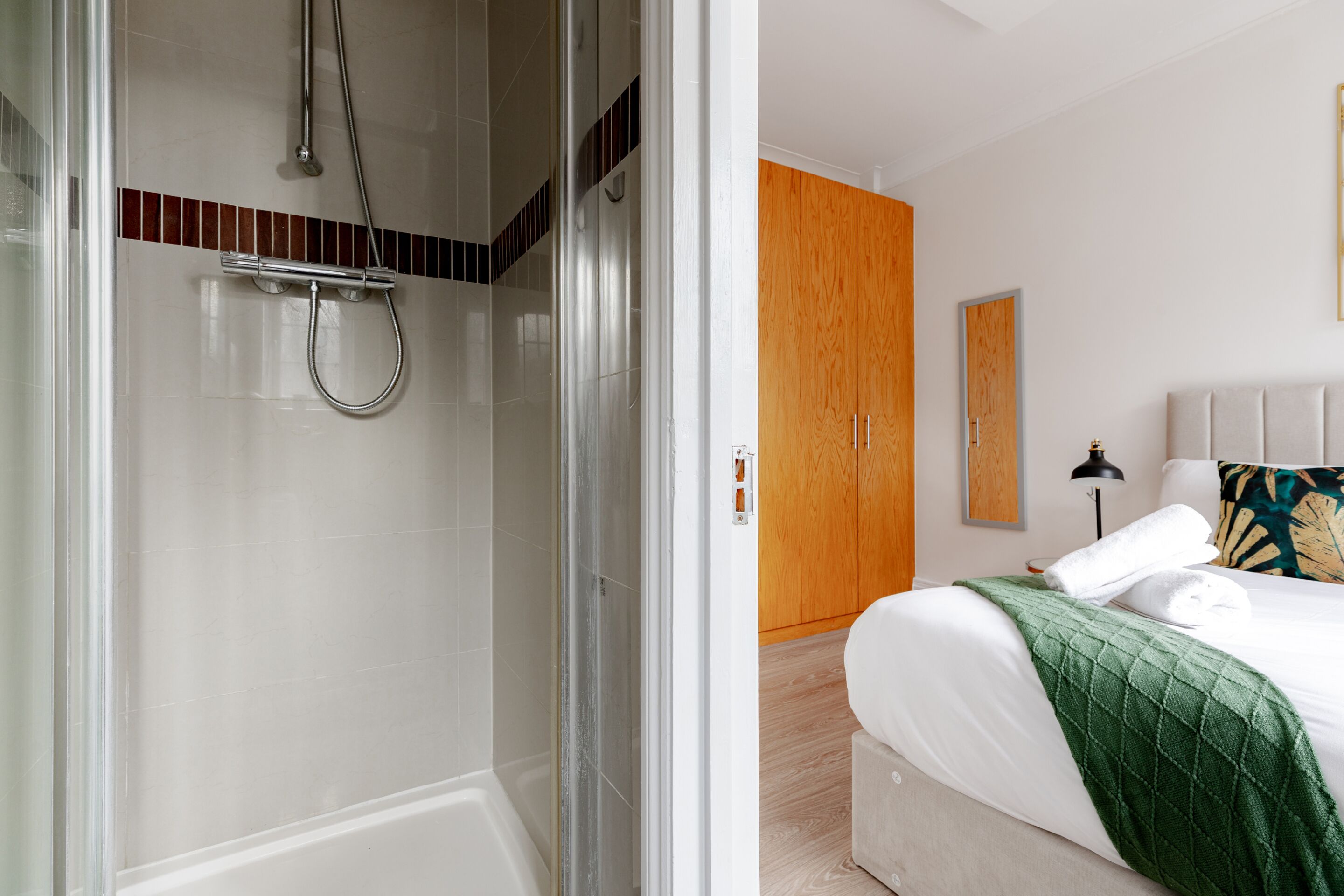 Une chambre d’hôtel moderne dotée d’une douche en verre, d’une armoire en bois, d’un lit recouvert d’une couverture verte et d’une table de chevet avec une lampe.