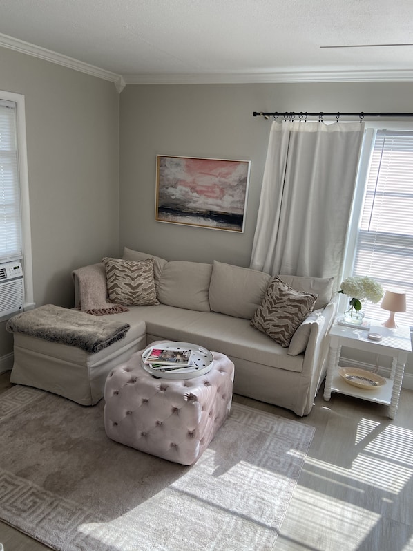 A living room with a beige sectional sofa, a tufted ottoman, a white side table, and a painting of a sunset. - Love Shack in Sherman (Sherman)