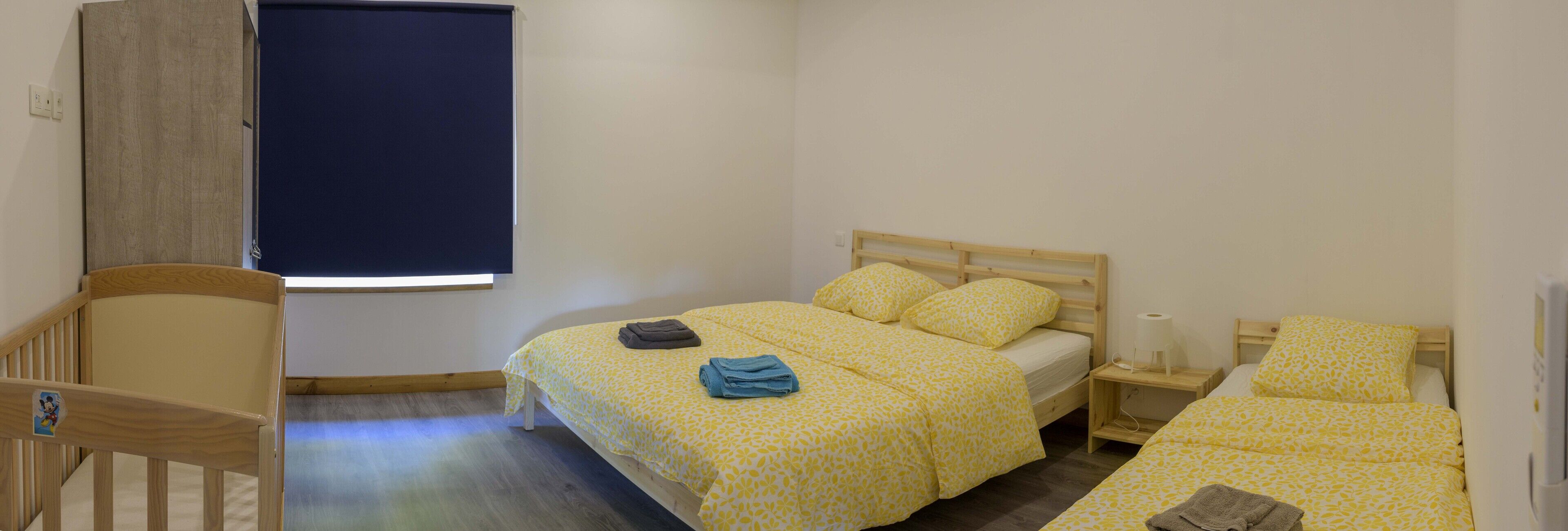 A bedroom with a single bed, a wooden crib, a bedside table with a lamp, and a window with a blind.