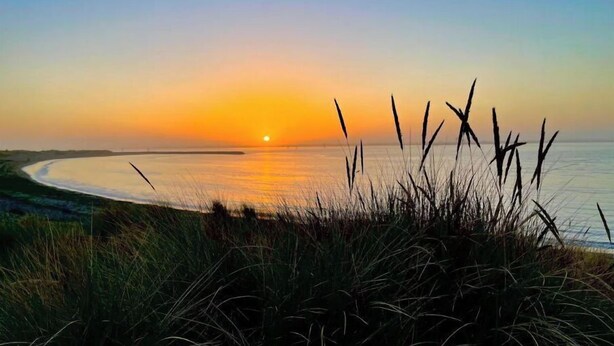 A beach scene with a sunset, grassy dunes, and a calm sea.