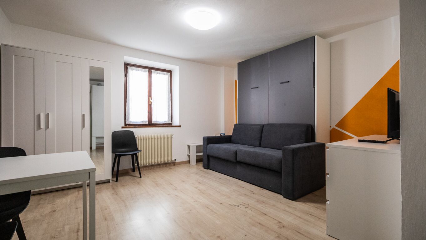 A modern, well-lit room with a grey sofa, a small table, a chair, and a TV on a cabinet.