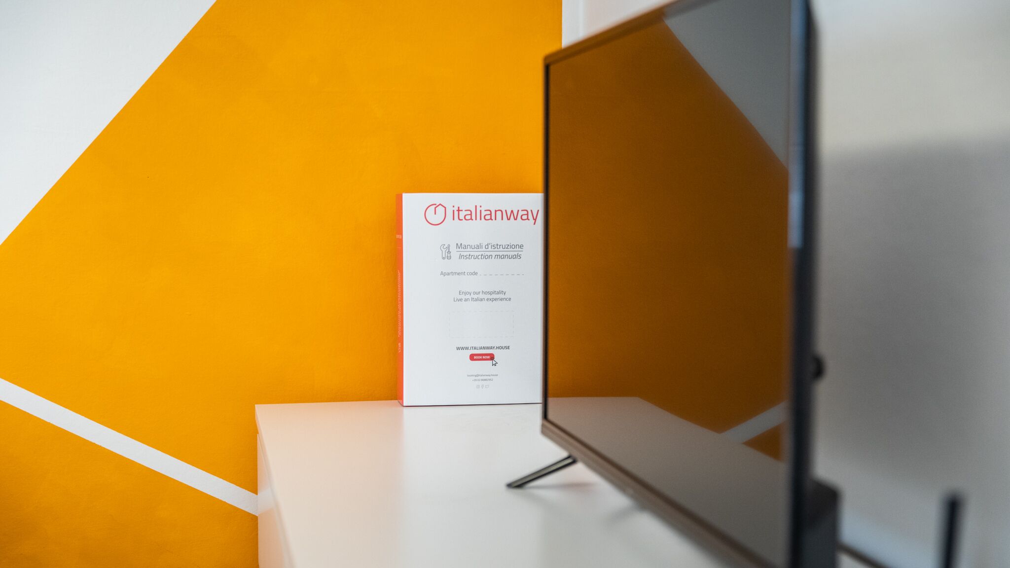 A sign for Italianway with a white background and red text, placed on a white surface against an orange wall.