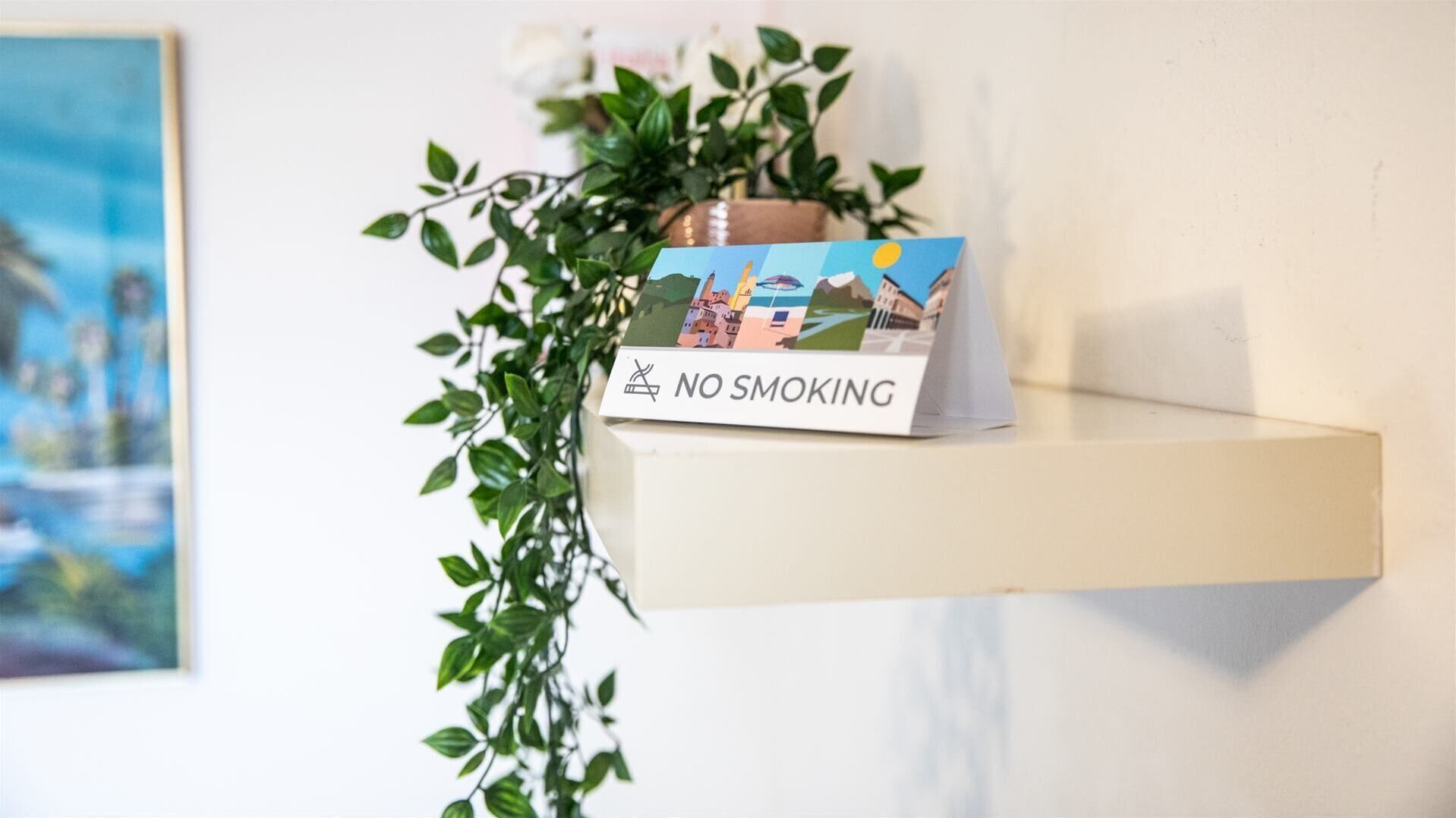A 'NO SMOKING' sign on a shelf with a plant and a postcard.
