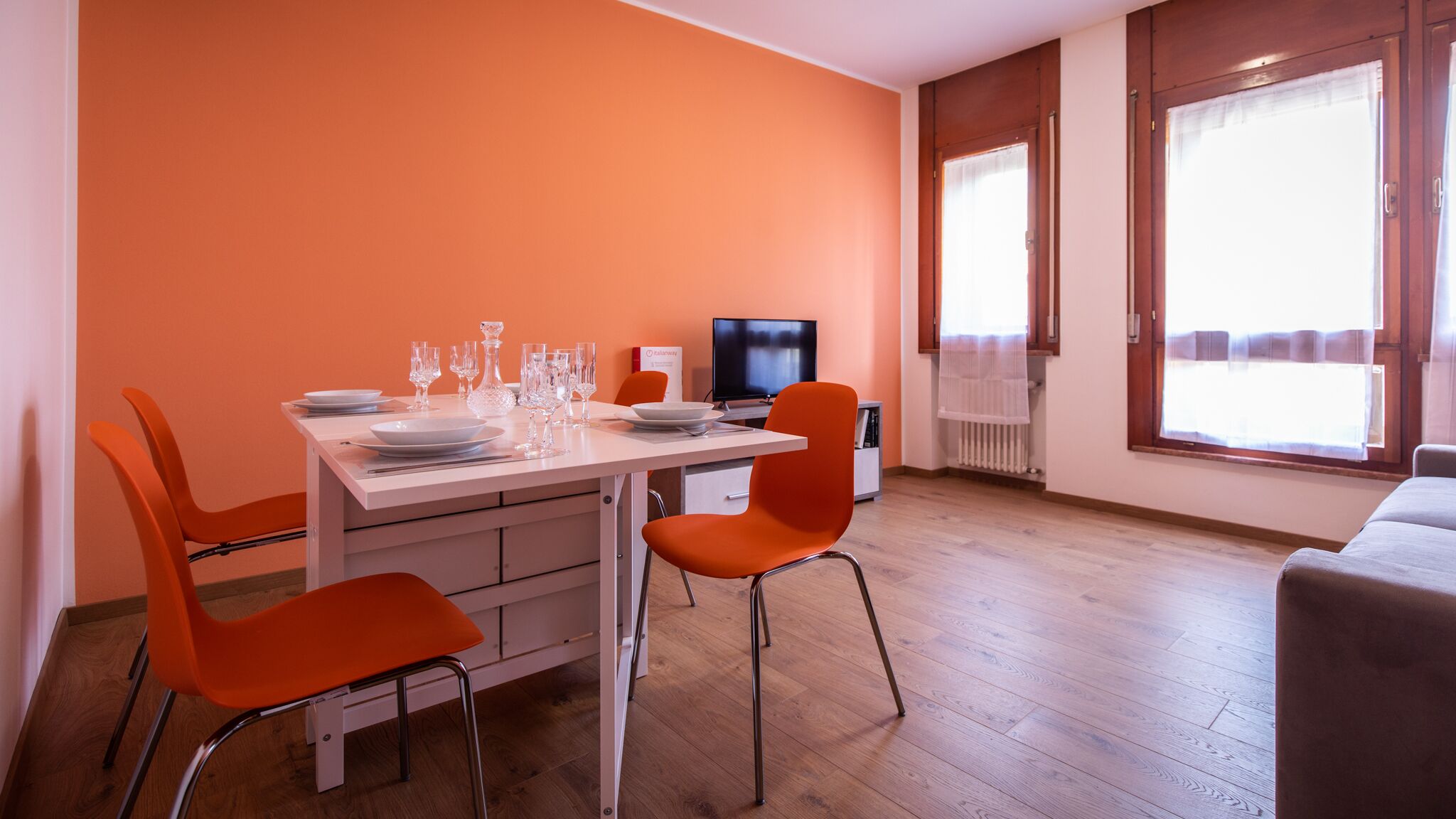 A dining area with a white table set for four, orange chairs, a television, and a window with curtains.