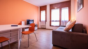 A modern living room with a white desk, orange chairs, a grey sofa, and large windows with curtains. - Italianway - Aquileia 53 (Udine)