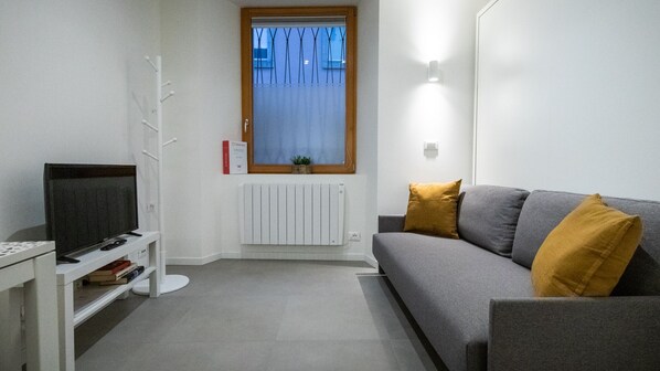 A modern living room with a grey sofa, a television on a stand, a radiator, and a window with blinds. - Italianway - Del Sale 4 (Udine)