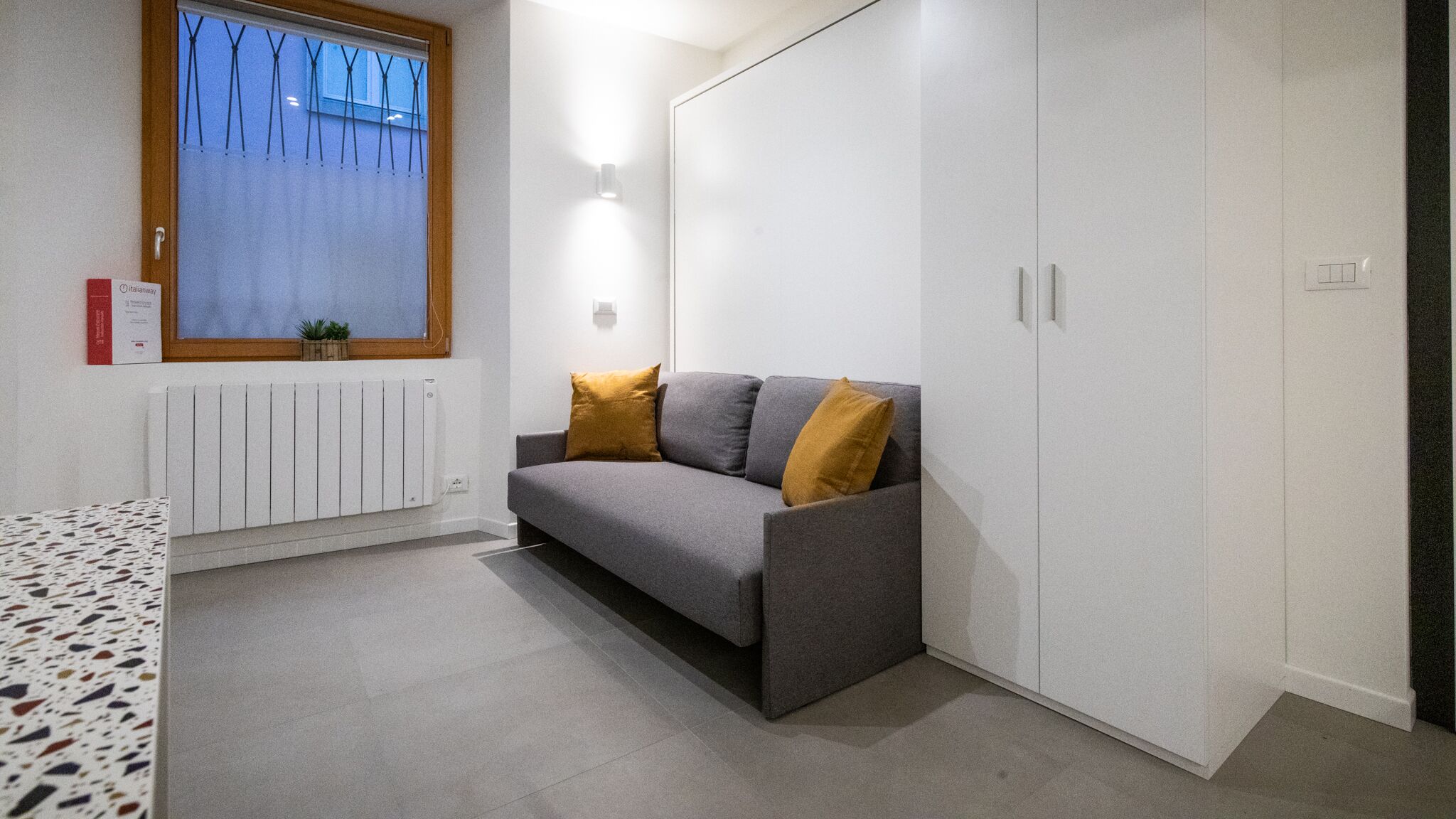 A modern, compact living space with a grey sofa, two mustard-colored cushions, a window with blinds, a radiator, and a wardrobe.