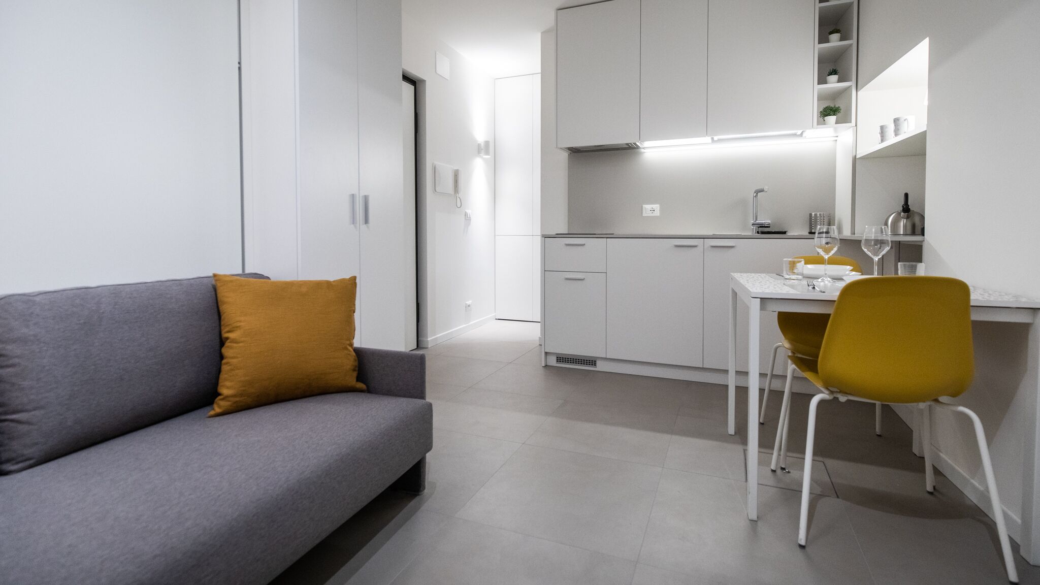 A modern, minimalist living space with a grey sofa, a mustard-colored pillow, a white dining table with a yellow chair, and a kitchen area with white cabinets and a kettle.