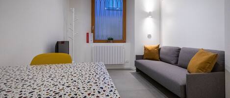 Una habitación pequeña y moderna con una cama, un sofá gris con dos cojines, una mesita y un radiador.