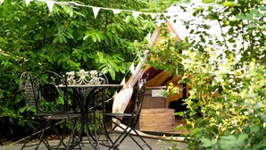 A rustic outdoor dining area with a metal table and chairs, surrounded by lush greenery and a wooden structure in the background. - Bedouin Bell Tent (Woolton Hill)