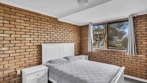 A bedroom with a brick wall, a white bed, a nightstand, and a window with curtains.