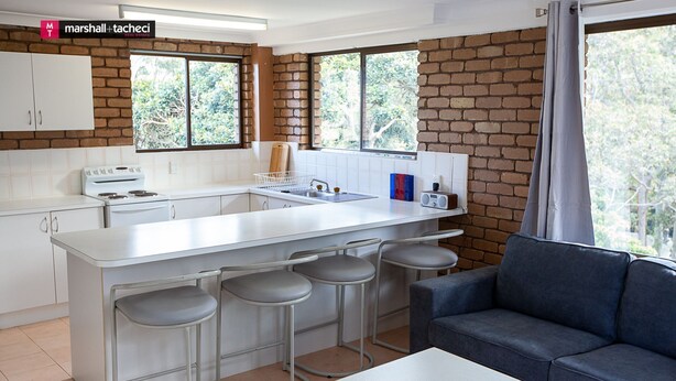 A modern kitchen with white cabinets, a central island with bar stools, and a large window with a view of trees. - George Hideaway - George St 30A (Bermagui)